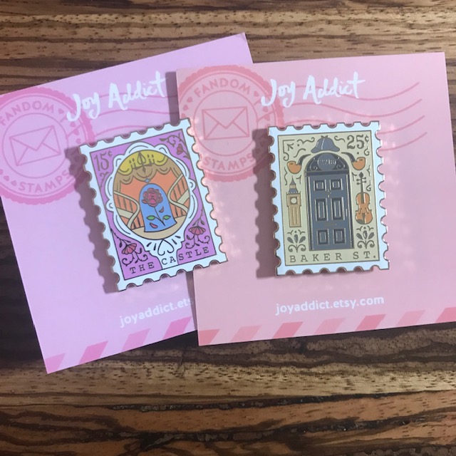 Stamp Enamel Pins on backing cards. One pin show a rose from beauty and the beast and the second shows the door of 221b baker street