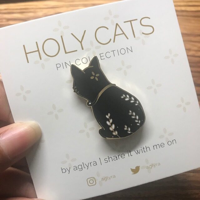 Enamel Pin featuring a black cat with leaves on its back on a white backing card which say holy cats