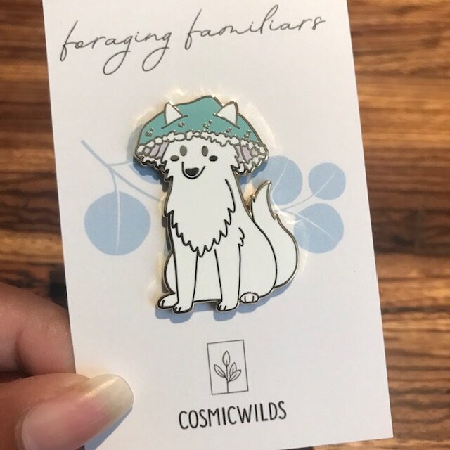 Enamel Pin of Arctic Wolf with a Verdigris Agaric Funghi on its head on a white backing card with the words Foraging Familiars