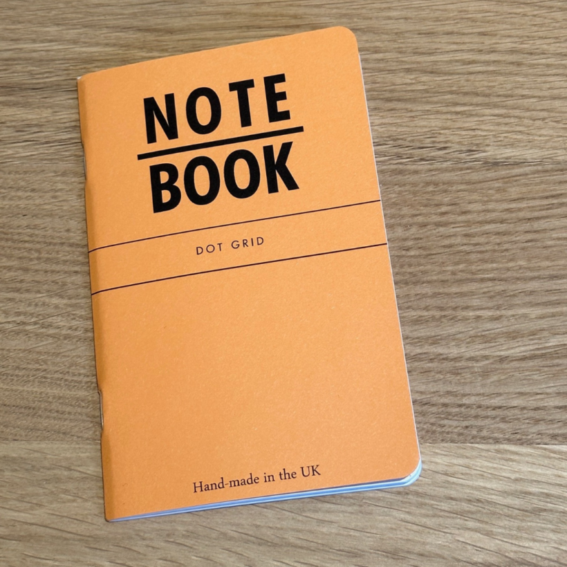orange A6 notebook with the words Note and the Book underneath in bold text and then dot grid in small print underneath.