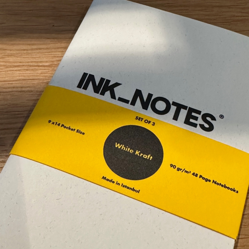 ocket notebook with a white kraft cover and the works "ink_notes" on the front cover wrapped in a bright yellow belly band with a black dot in the middle with the words "white kraft"