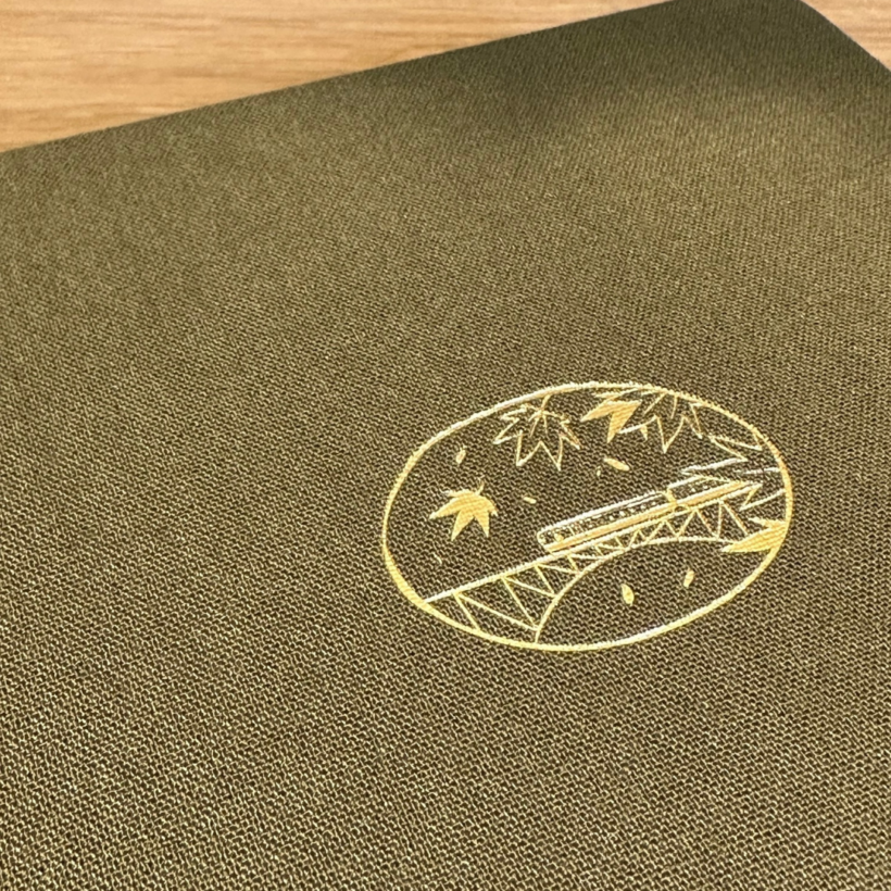 bullet journal with gold foil illustration of bullet train on bridge with leaves and an olive green linen cover