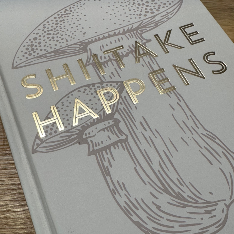 mushroom coloured hard cover notebook with an image of a shitake mushroom and the words "shitake Happens" in gold on the cover.