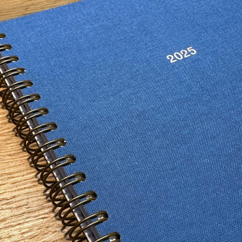 The image shows a close-up of a spiral-bound planner with a blue fabric cover and "2025" embossed in white on the front. It is resting on a wooden surface.