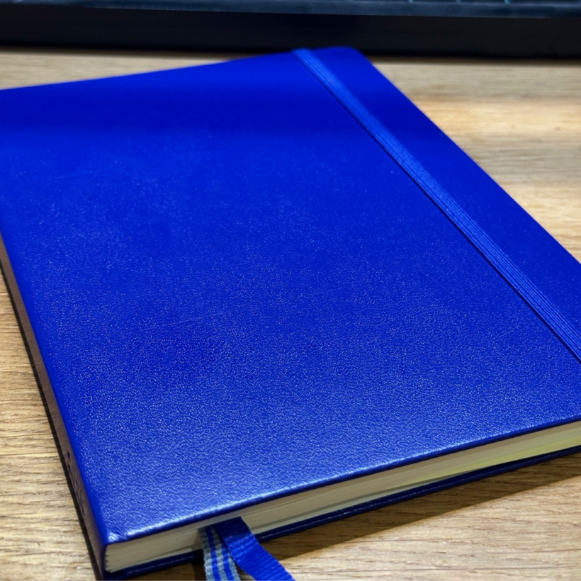 A closed notebook with a bright blue textured cover, featuring an elastic band on the right side and a striped blue ribbon bookmark peeking out. The notebook is placed on a wooden surface with a keyboard visible in the background.