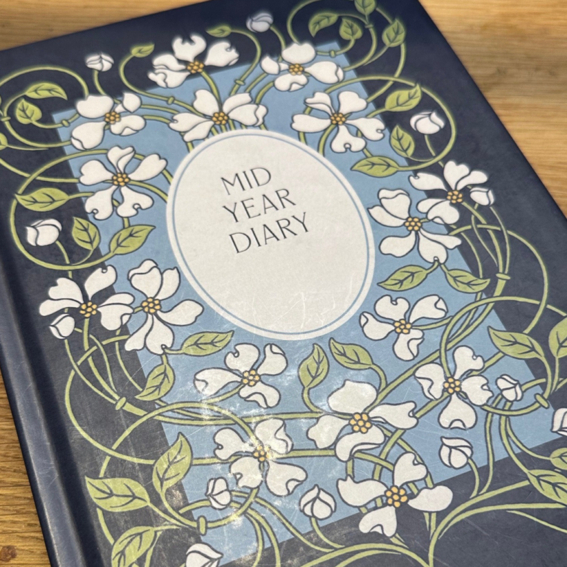 A decorative diary titled "Mid Year Diary" with a floral design on the cover. The design features white flowers and green vines on a blue and black background. The diary is resting on a wooden surface.