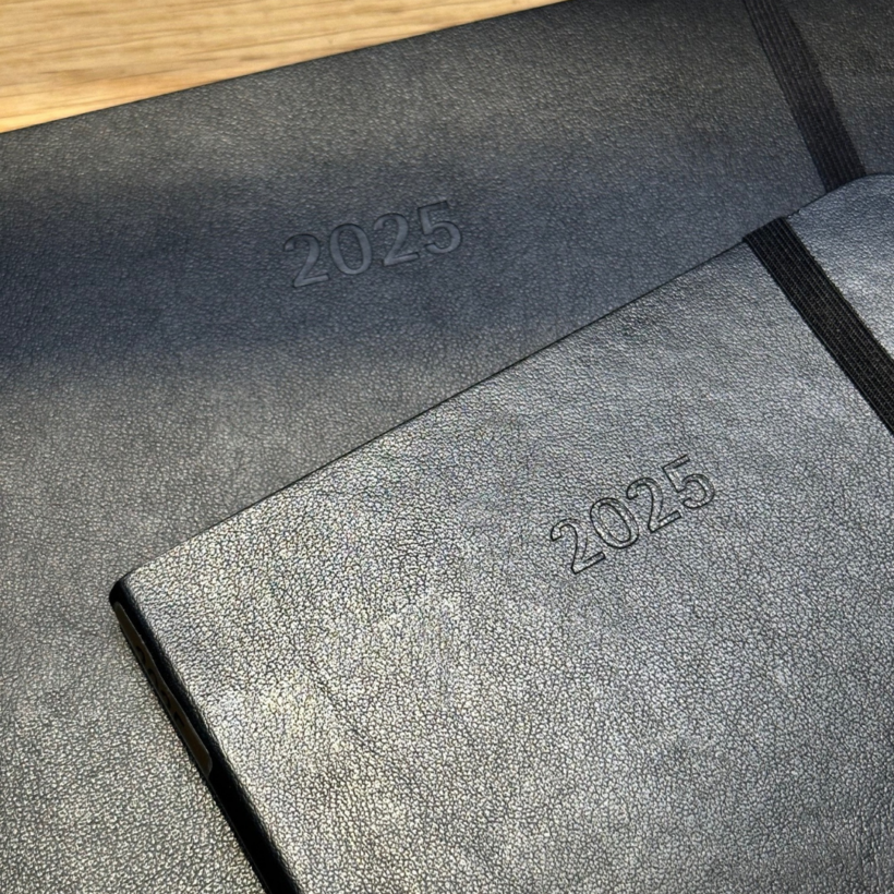 Two black 2025 planners with textured covers are placed on a wooden surface. One planner is slightly atop the other, with an elastic band visible on the top-right corner of the closer planner. Both have "2025" embossed on the cover.