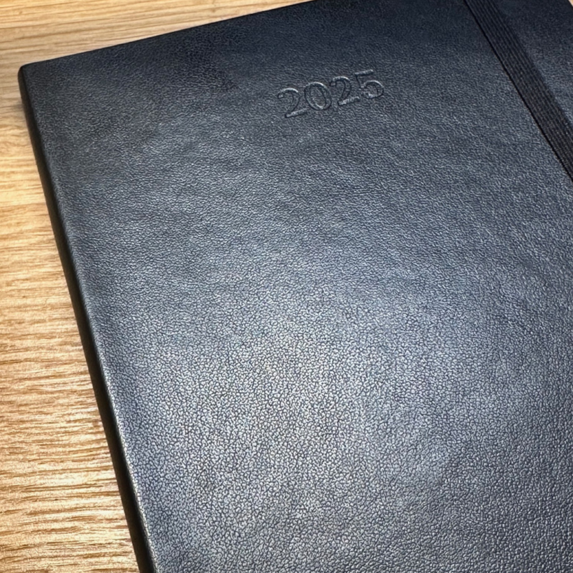 A textured black notebook with "2025" embossed on the cover. It features an elastic band on the right side and is placed on a wooden surface.