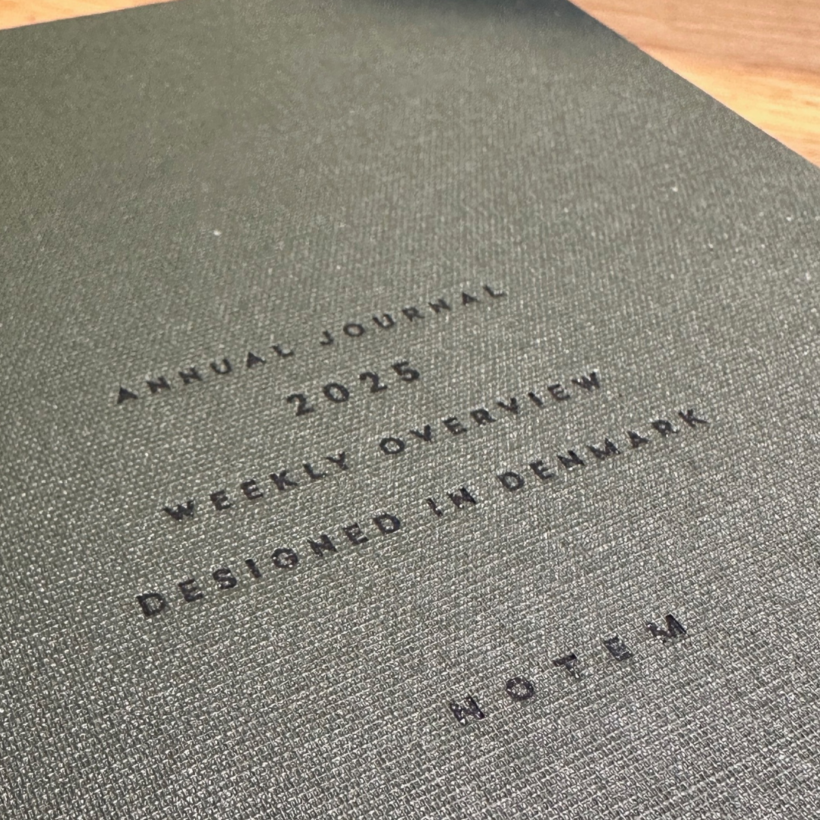 A close-up of a textured gray cover of an annual journal. The text, embossed in black, reads: "ANNUAL JOURNAL 2025 WEEKLY OVERVIEW DESIGNED IN DENMARK NOTEM."