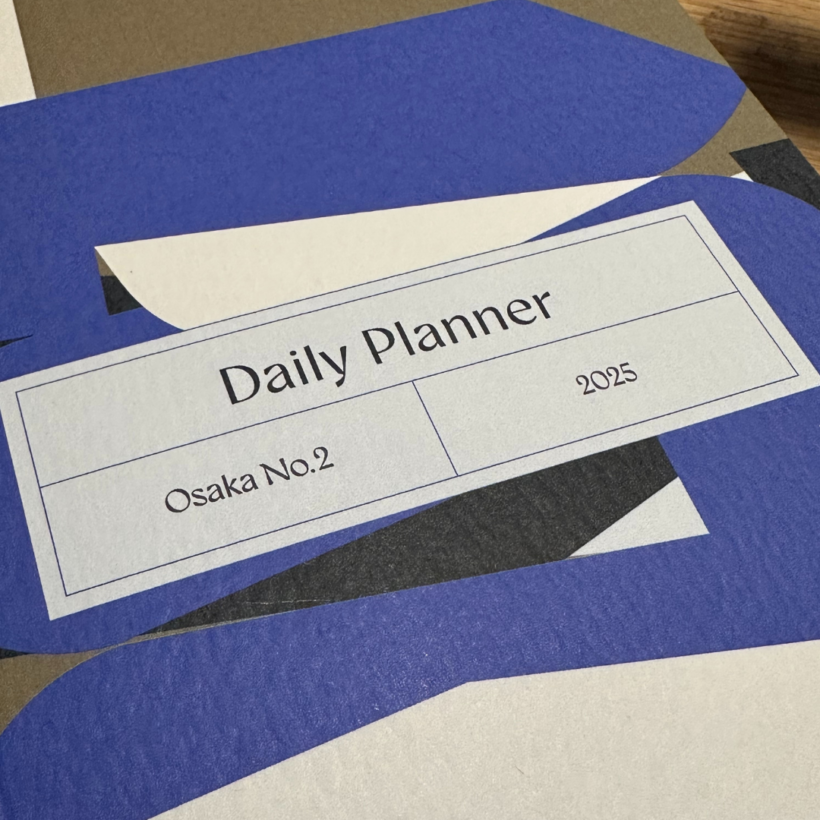 A close-up of a 2025 daily planner titled "Osaka No.2." The cover features a minimalist design with overlapping blue, white, black, and brown geometric shapes. The title is printed in a clean font within a rectangular frame.