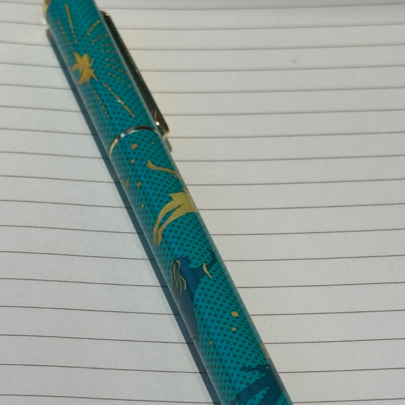 A close-up of a teal pen with a gold design, including stars and silhouettes, resting on a lined notebook. The background shows blank lined pages.